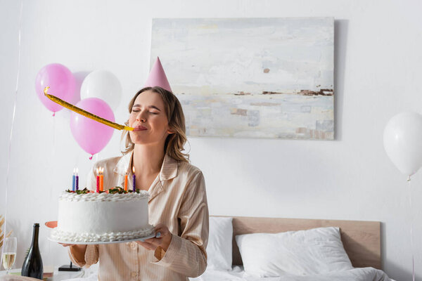 Woman blowing party horn and holding birthday cake on bed 