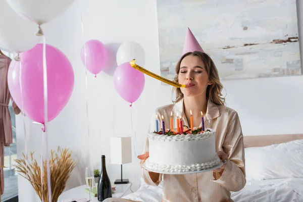 Woman with party horn and cap holding birthday cake near balloons in bedroom
