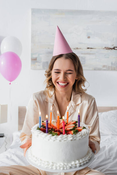 Positive woman in party cap holding birthday cake with candles in bedroom 