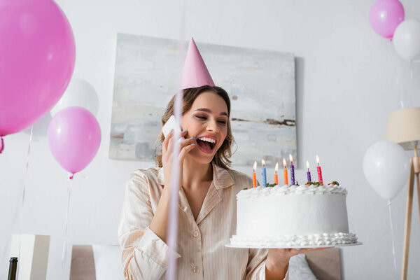 Woman with birthday cake talking on smartphone and holding birthday cake near balloons at home 