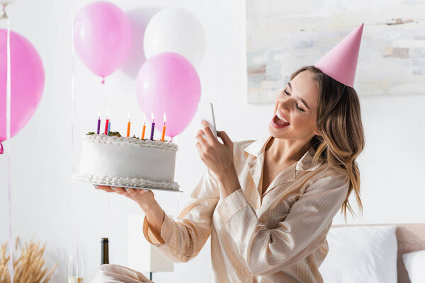 Cheerful woman with birthday cake taking photo on smartphone during party at home 