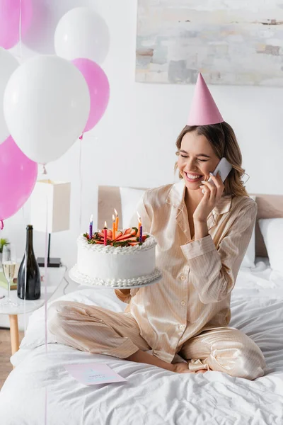 陽気な女性は携帯電話で話をし ベッドルームでお祝いの間に誕生日ケーキを保持 — ストック写真