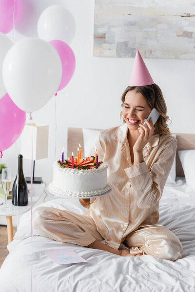 Cheerful woman talking on mobile phone and holding birthday cake during celebration in bedroom 