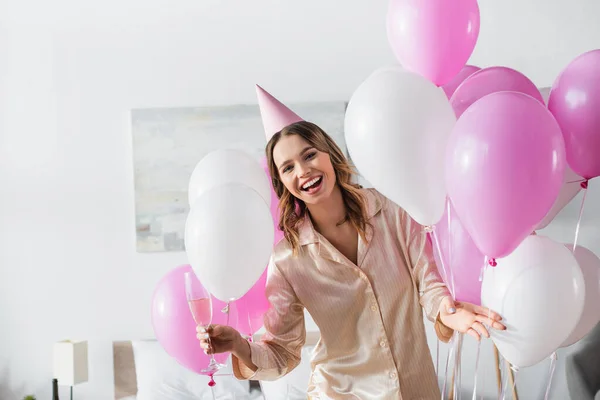 Cheerful woman in pajama and party cap holding glass of champagne near balloons in bedroom