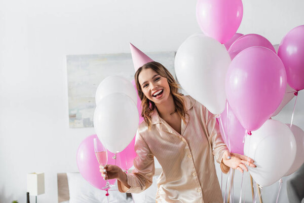 Cheerful woman in pajama and party cap holding glass of champagne near balloons in bedroom 