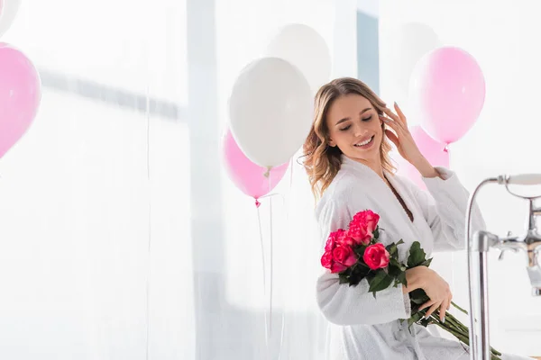 Cheerful woman holding roses near balloons in bathroom