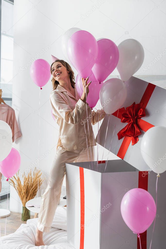 Young woman opening huge present with festive balloons during birthday party in bedroom 