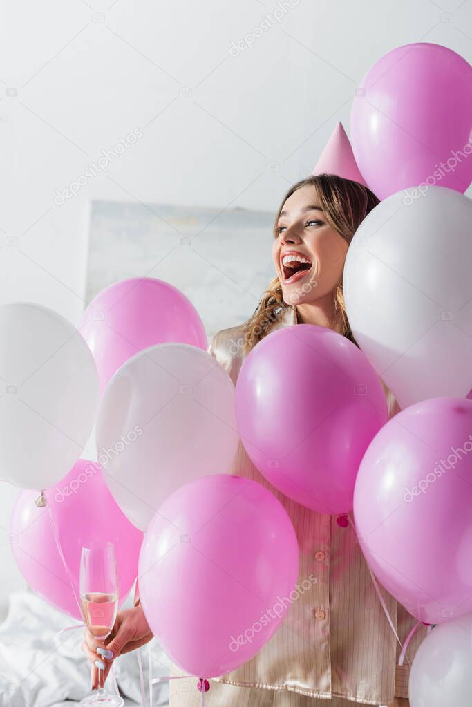 Excited woman with champagne looking away near festive balloons in bedroom 