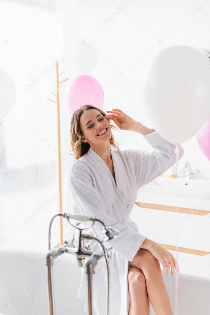 Happy woman in bathrobe sitting on bathtub near blurred balloons 