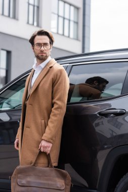 young man in glasses and coat standing with briefcase near car 