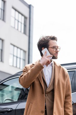 businessman in glasses and coat talking on phone near car 