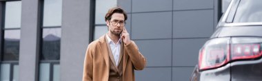 confident businessman in glasses and beige coat talking on phone near car, banner
