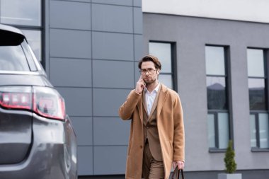 confident businessman in glasses and beige coat talking on mobile phone near car
