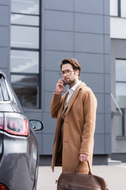 elegant businessman in glasses and beige coat talking on mobile phone near car