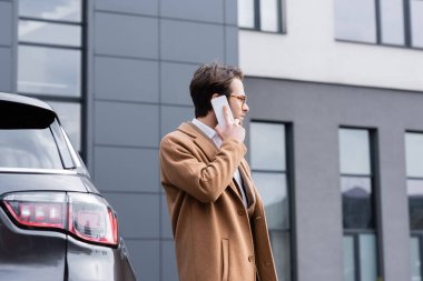 side view of confident man in glasses and beige coat talking on mobile phone near car