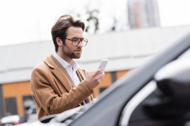 confident man in glasses and beige coat looking at mobile phone near blurred car