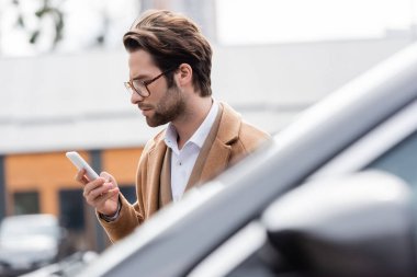 young man in glasses and beige coat looking at cellphone near blurred car