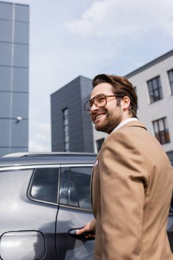happy young businessman in beige jacket smiling near modern car 