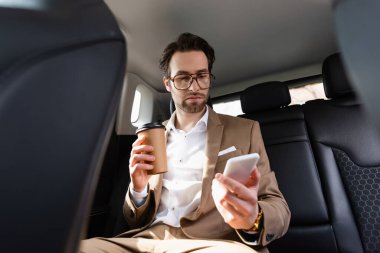 man in suit and glasses holding paper cup and looking at smartphone in car 