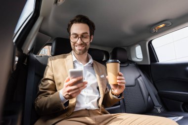 joyful businessman in suit and glasses holding paper cup and smartphone in car 