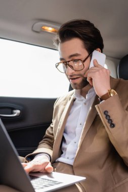 businessman working remotely with laptop while talking on smartphone in car