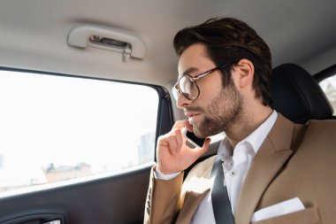 bearded man in glasses and suit talking on smartphone in car
