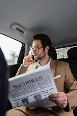 confident man in glasses talking on smartphone and holding business newspaper in car 