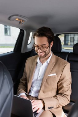 cheerful businessman in glasses typing on laptop in car