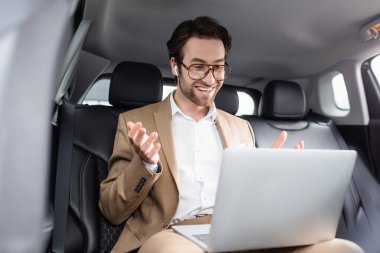smiling businessman in glasses and wireless earphones gesturing during video call in car 