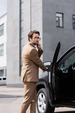 man in suit and glasses talking on smartphone and standing near car with opened door