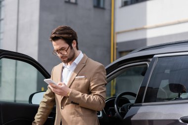 bearded man in suit and glasses looking at cellphone and standing near car with opened door