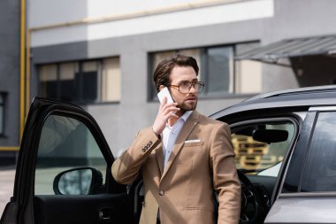 bearded businessman in suit and glasses talking on cellphone and standing near car with opened door