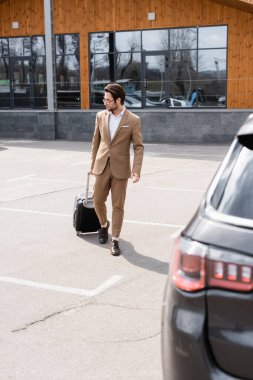 full length of man in suit and glasses walking with suitcase near car on parking 