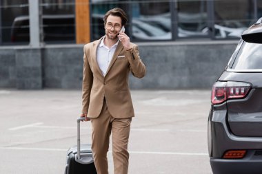 cheerful man in suit and glasses talking on smartphone while walking near car with suitcase