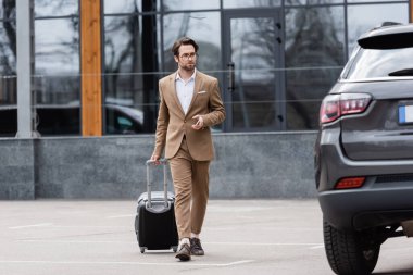 full length of man in suit and glasses walking near car with suitcase 