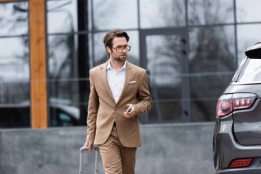 man in beige suit and glasses walking near car with suitcase 