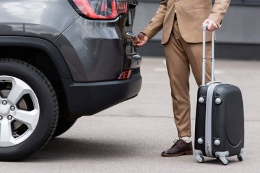 partial view of man in suit standing with suitcase and opening car trunk 