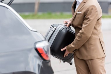 cropped view of man in suit putting suitcase in car trunk 