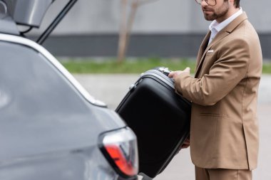 cropped view of bearded man in suit putting suitcase in car trunk 