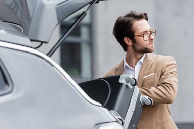 distracted man in glasses and suit putting baggage in blurred car trunk  clipart