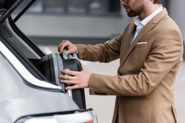 cropped view of bearded man in suit putting luggage in car trunk 