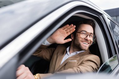 happy man in glasses looking through car window and waving hand while driving auto 