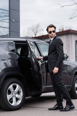 bearded bodyguard in suit and sunglasses with security earpiece opening car door 