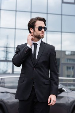 bearded bodyguard in suit and sunglasses with security earpiece near modern building on blurred background 