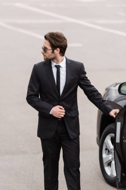 bearded bodyguard in suit and sunglasses with security earpiece opening door of modern auto 