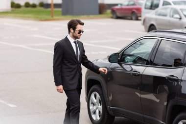 bearded bodyguard in suit and sunglasses with security earpiece walking near modern car