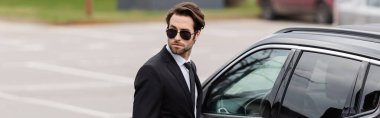 bearded bodyguard in suit and sunglasses with security earpiece near modern auto, banner