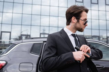 bearded bodyguard in sunglasses and suit holding gun near modern building 