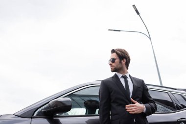 low angle view of bearded bodyguard in sunglasses and suit standing near modern car