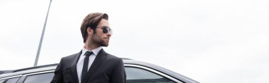low angle view of bearded bodyguard in sunglasses and suit standing near modern car, banner